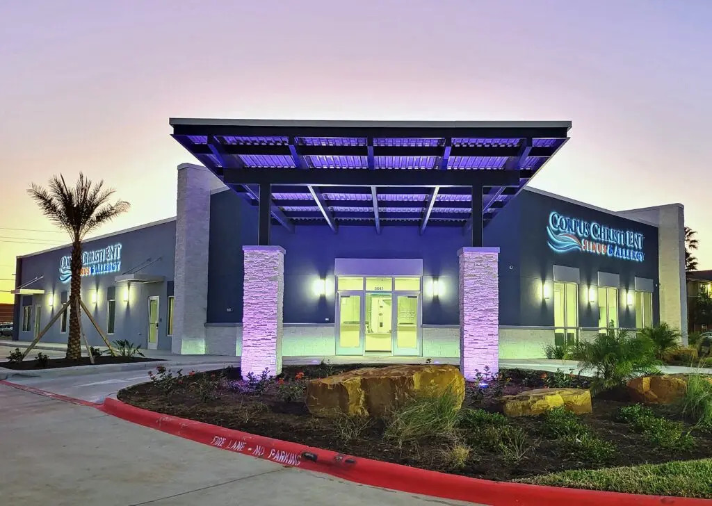 A modern building with a covered entrance, glowing purple lights, and landscaped surroundings at sunset. The sign reads Coastal Children’s Bay Ambulatory Surgery. Palm trees and rocks decorate the exterior.